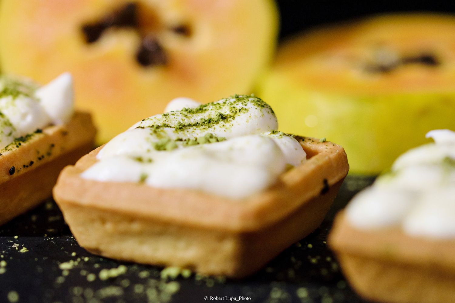 Mini tart shells topped with whipped cream and green zest, served alongside fresh sliced papaya on a dark surface.