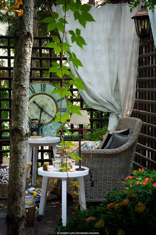 Cozy garden patio with wicker chair, white side tables, large clock, sheer curtains, and lush green plants.