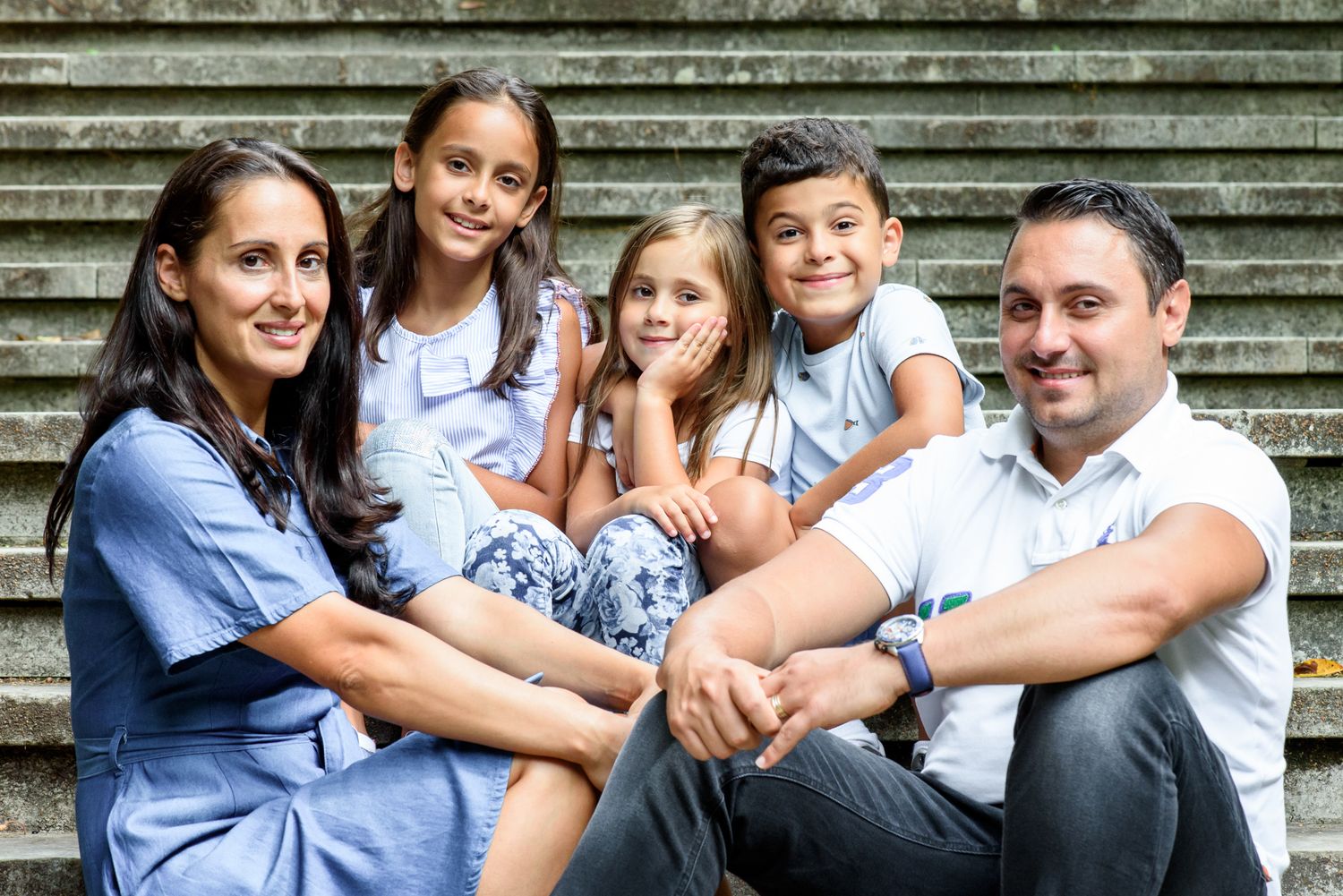 Happy family of five sitting on stone steps, smiling together outdoors in casual blue outfits.