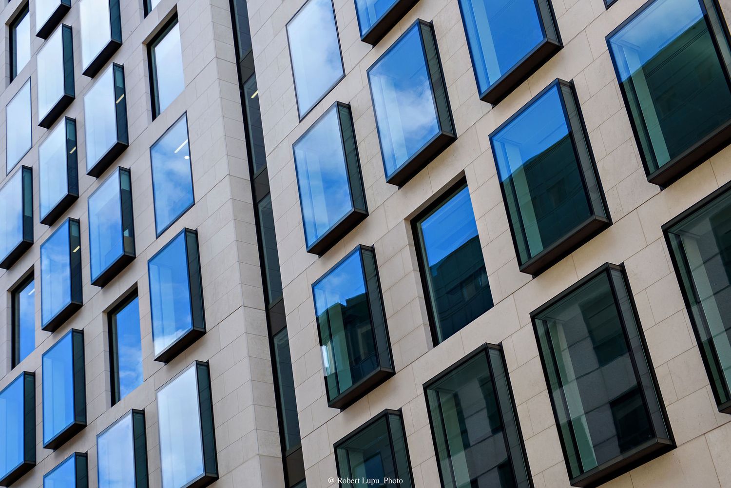 Modern office building facade with blue reflective windows mirroring cloudy sky, geometric pattern architecture.