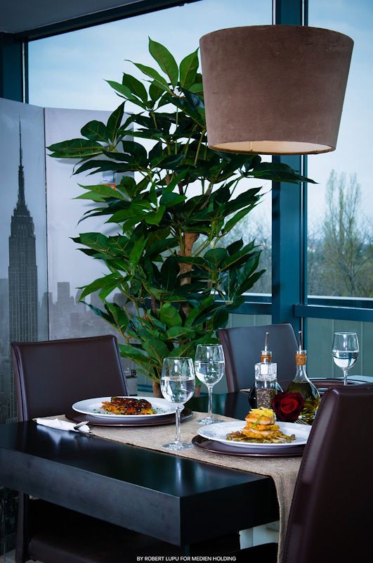 Elegant restaurant table set for two with gourmet dishes, wine glasses, and a lush plant near a city-view window.