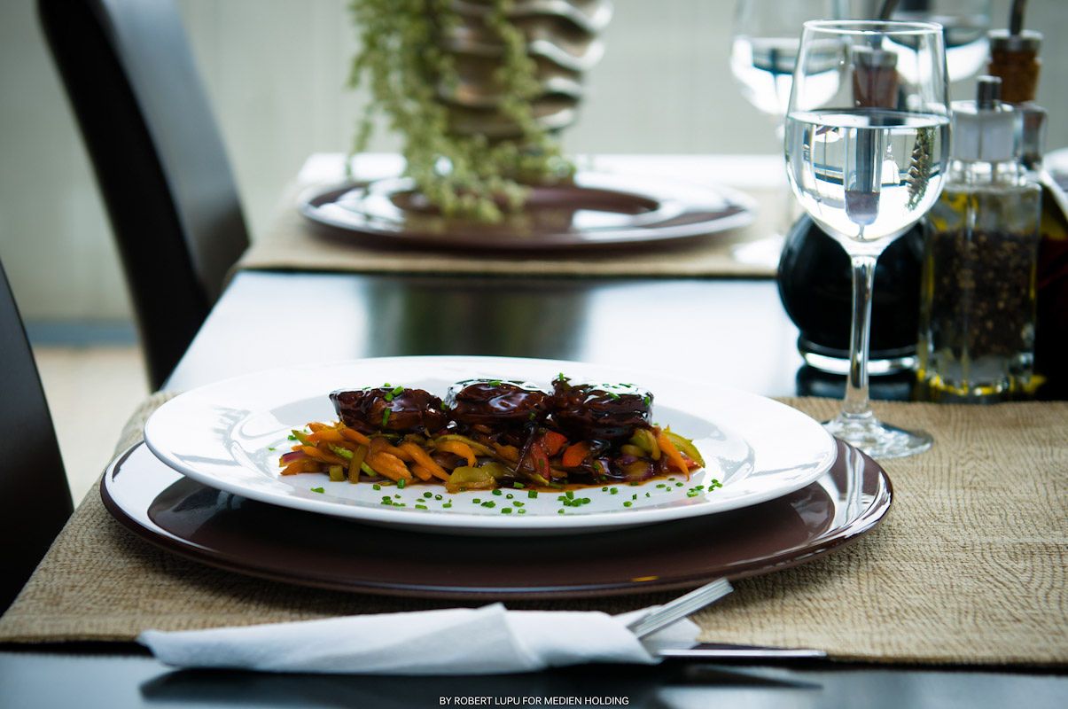 Elegant restaurant plate with braised meat on colorful vegetables, served on white dish with wine glass in background.