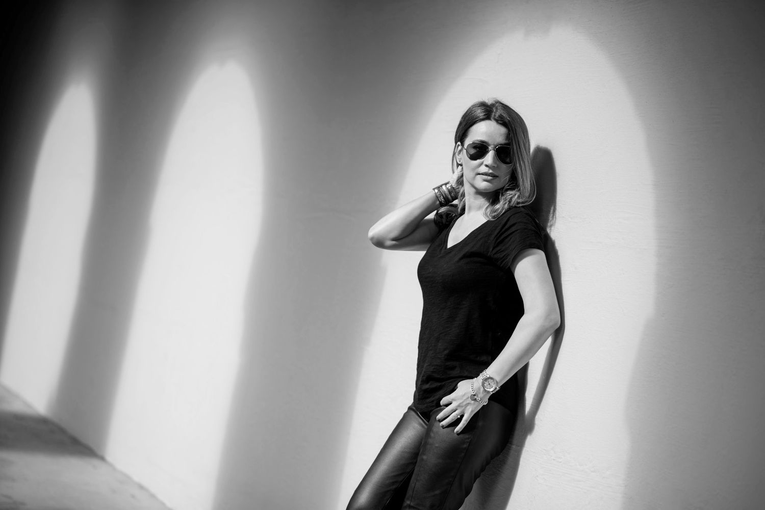 Black and white photo of a stylish woman in sunglasses and black outfit leaning against a wall with circular shadows.