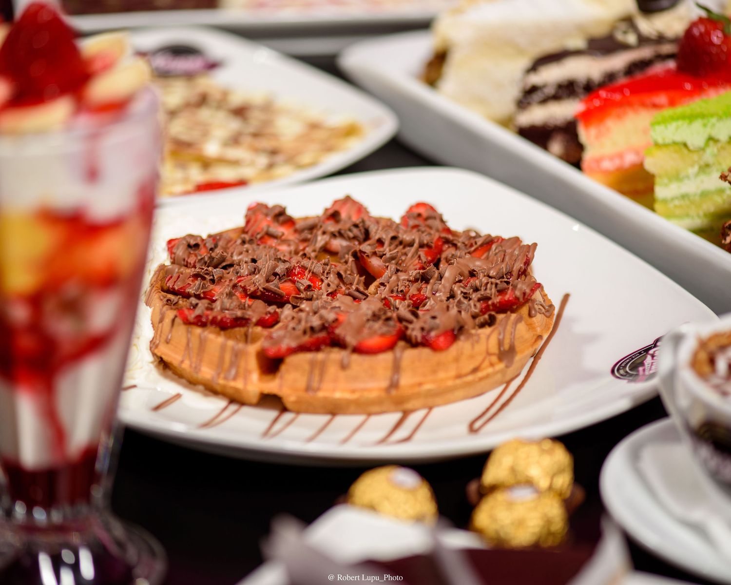 Chocolate waffle topped with red berries and brownie pieces on a white plate, surrounded by desserts.