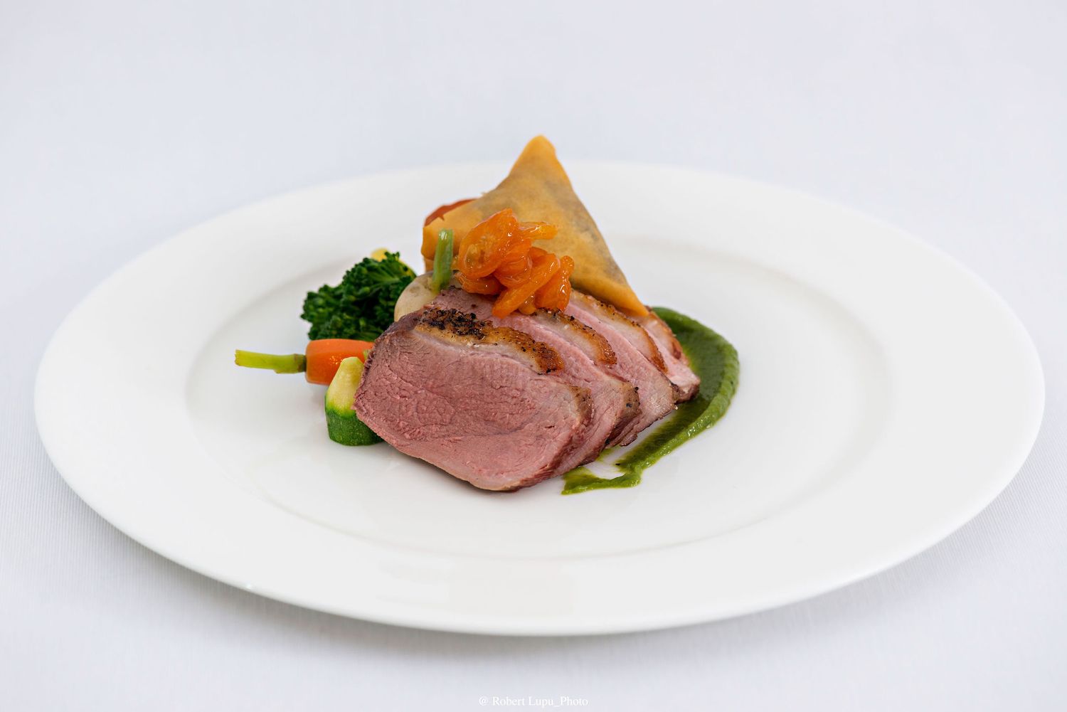 Elegant plated duck breast with green herb sauce, roasted vegetables, and a crispy pastry garnish on a white plate.