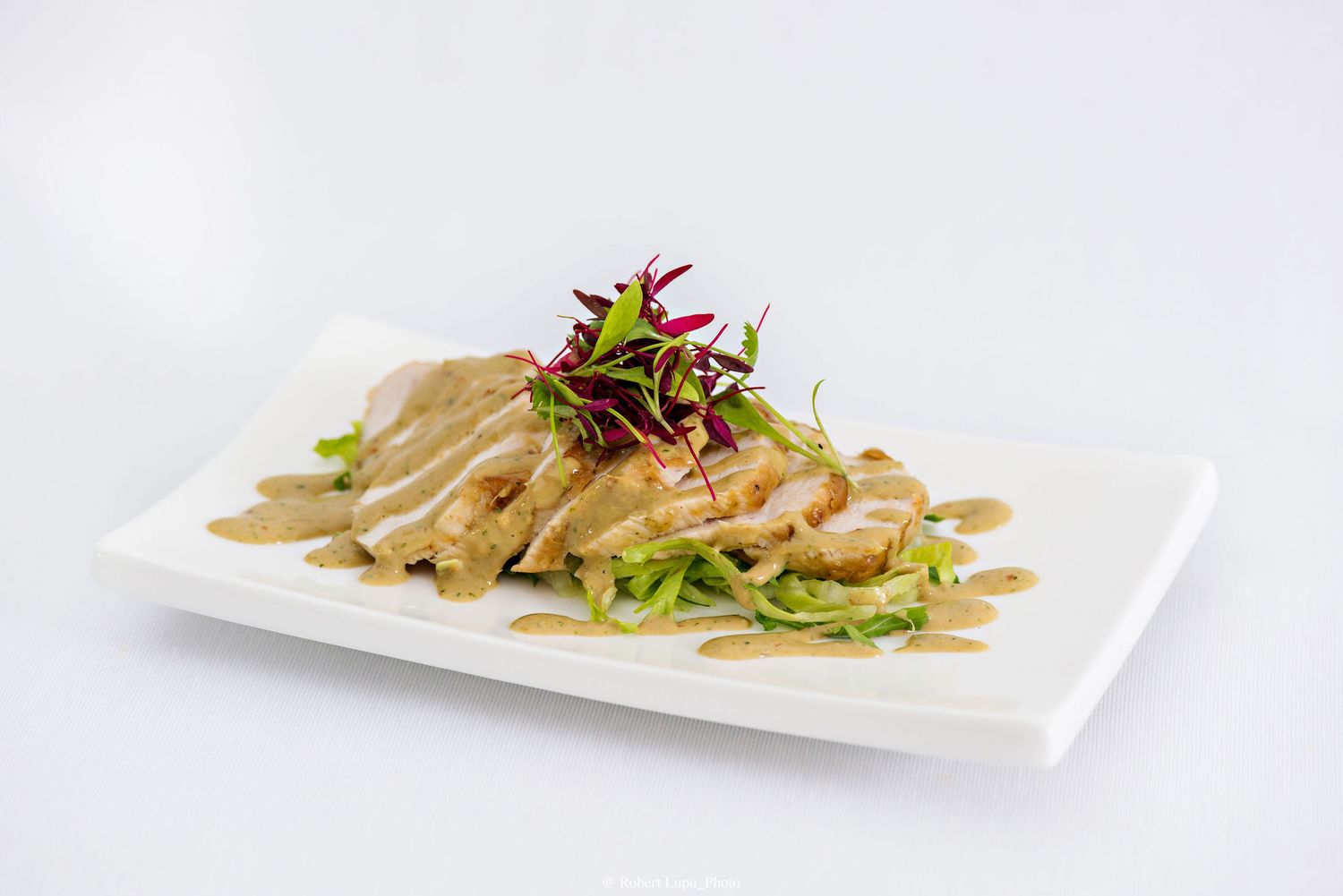 Sliced chicken breast with creamy mushroom sauce, fresh greens, and microgreens garnish on a white plate.