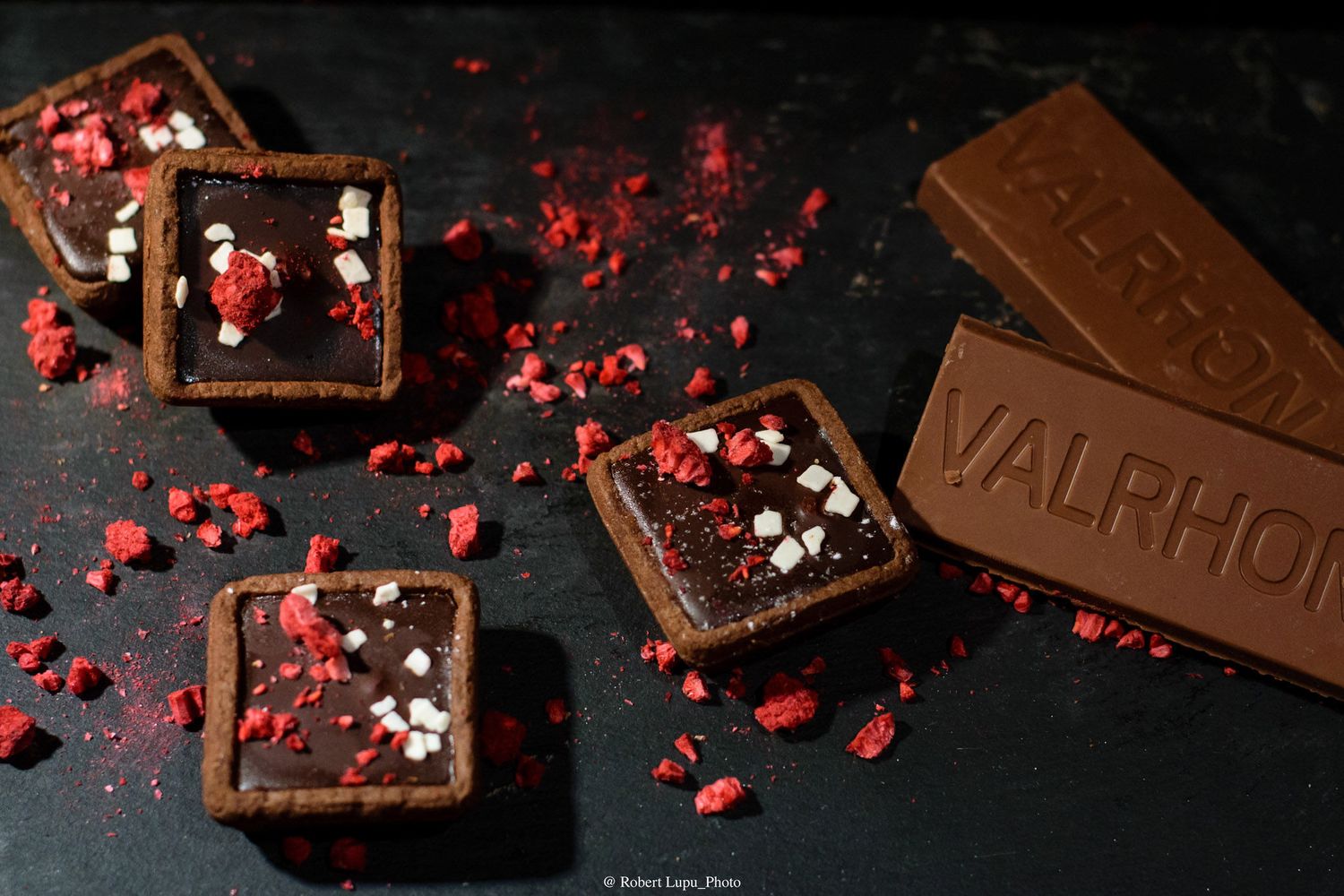Square chocolate tarts topped with freeze-dried raspberries beside a Valrhona chocolate bar on dark background.