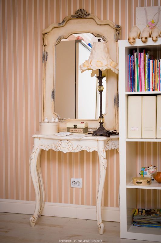 Elegant vintage white vanity table with ornate mirror, lamp, and bookshelf in a pink striped wallpaper room.