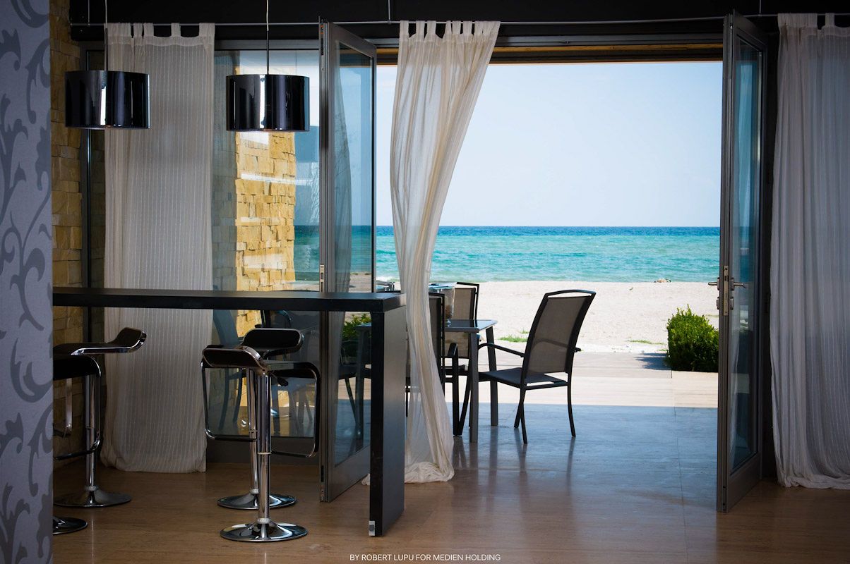 Elegant beachfront bar with black stools and white curtains opening to a stunning ocean view.