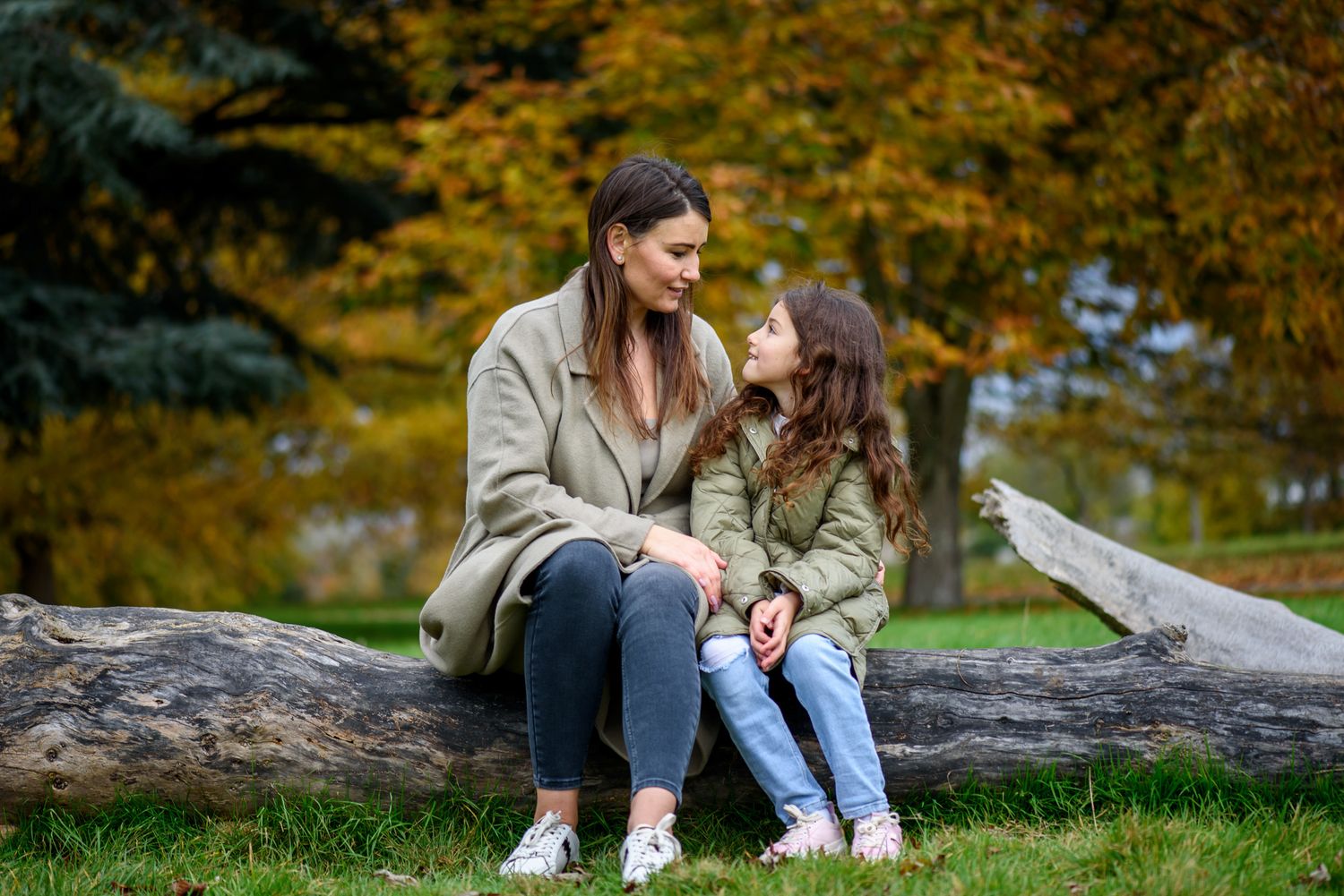 Mother and daughter sitting on a fallen log in an autumn park, smiling at each other surrounded by colourful foliage.