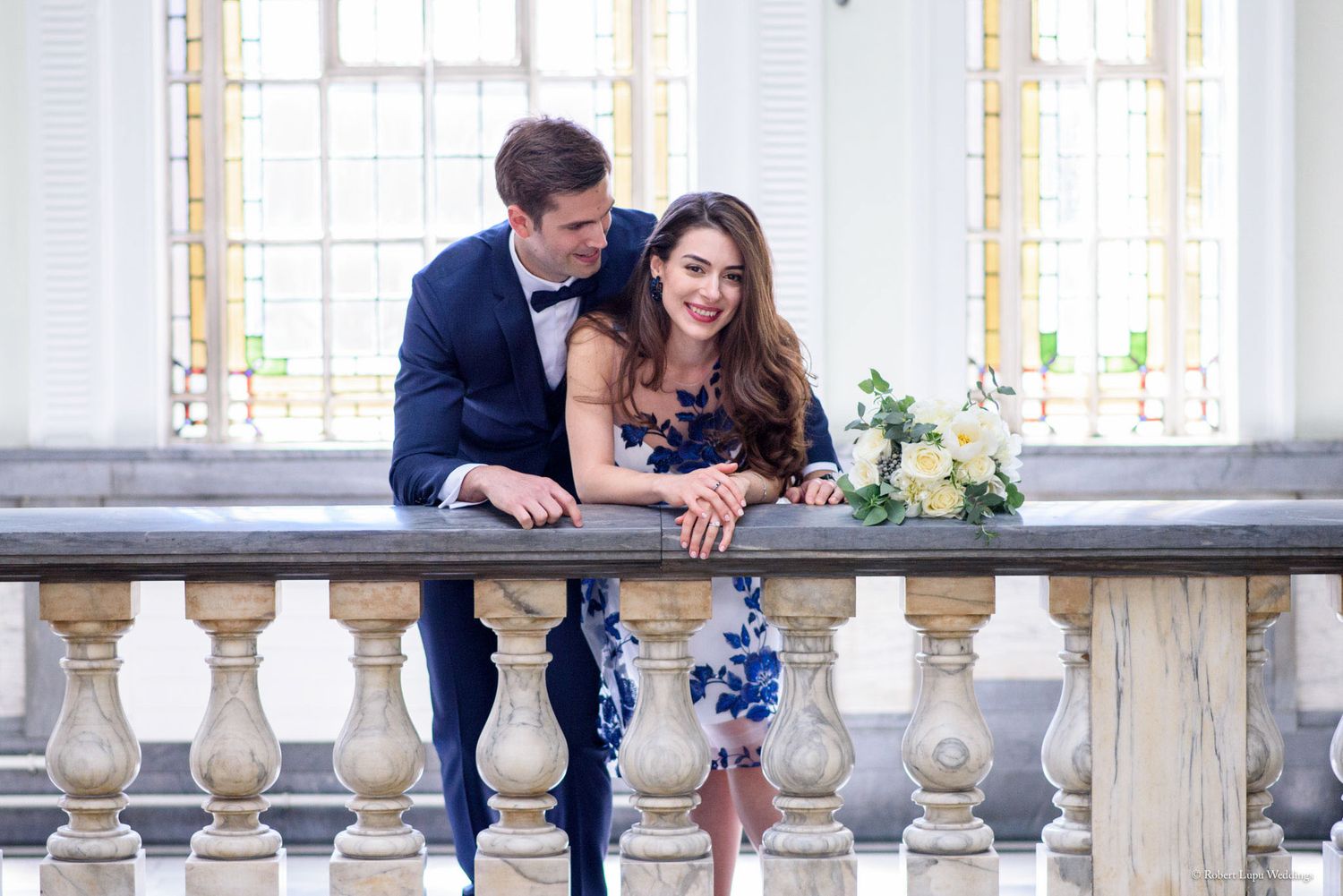 Elegant couple posing on a balcony, man in navy tuxedo and woman in blue floral dress holding a bouquet.