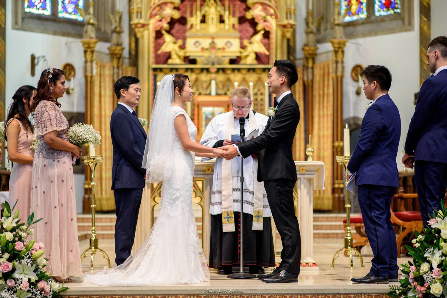 Couple exchanging vows at a church wedding ceremony with officiant and wedding party at ornate altar.