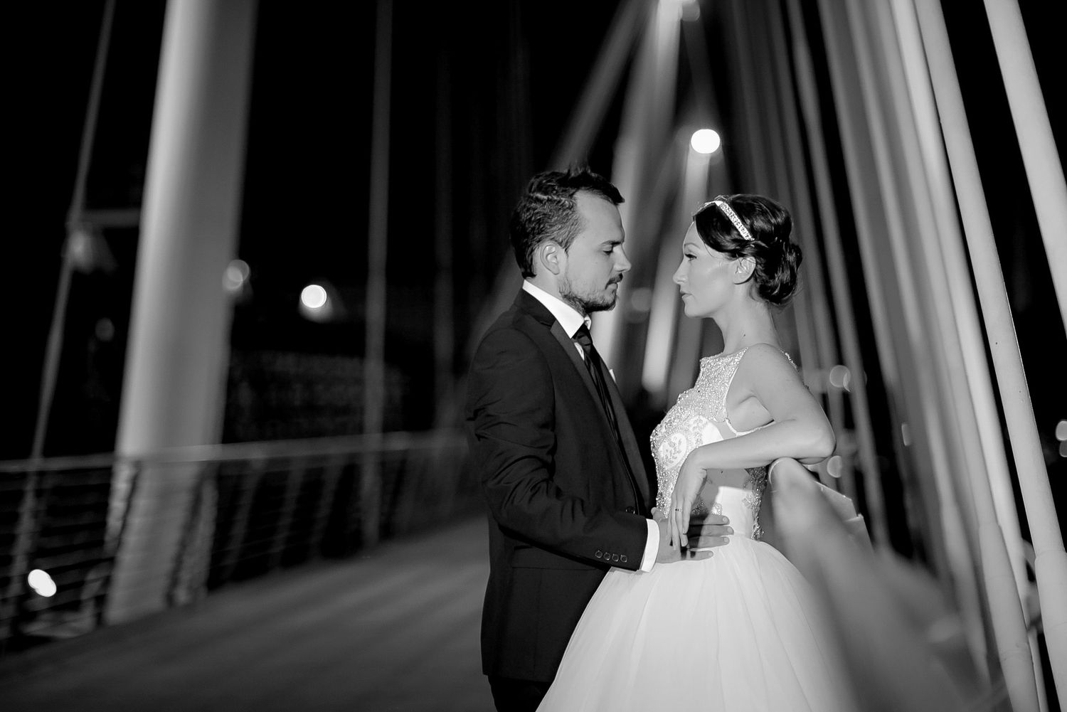 Black and white wedding photo of bride and groom sharing a romantic moment on a bridge at night.