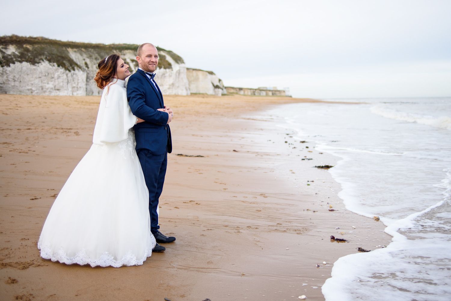Bride and groom standing close together on a sandy beach with white chalk cliffs behind.