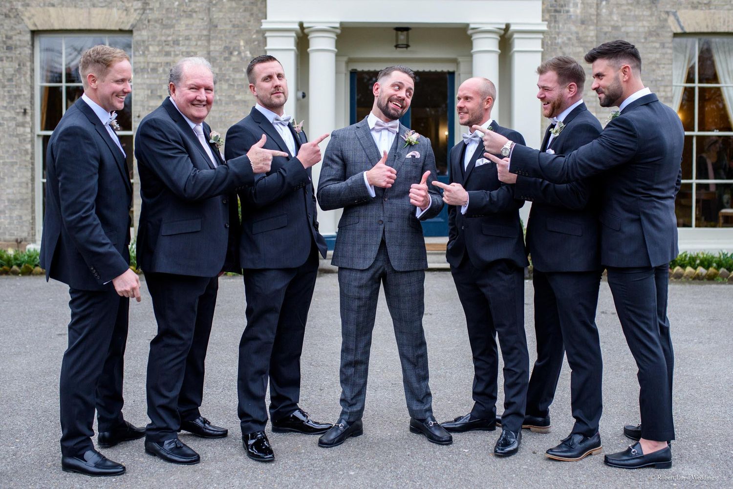 Groom and groomsmen in dark suits laughing and pointing outside an elegant wedding venue.