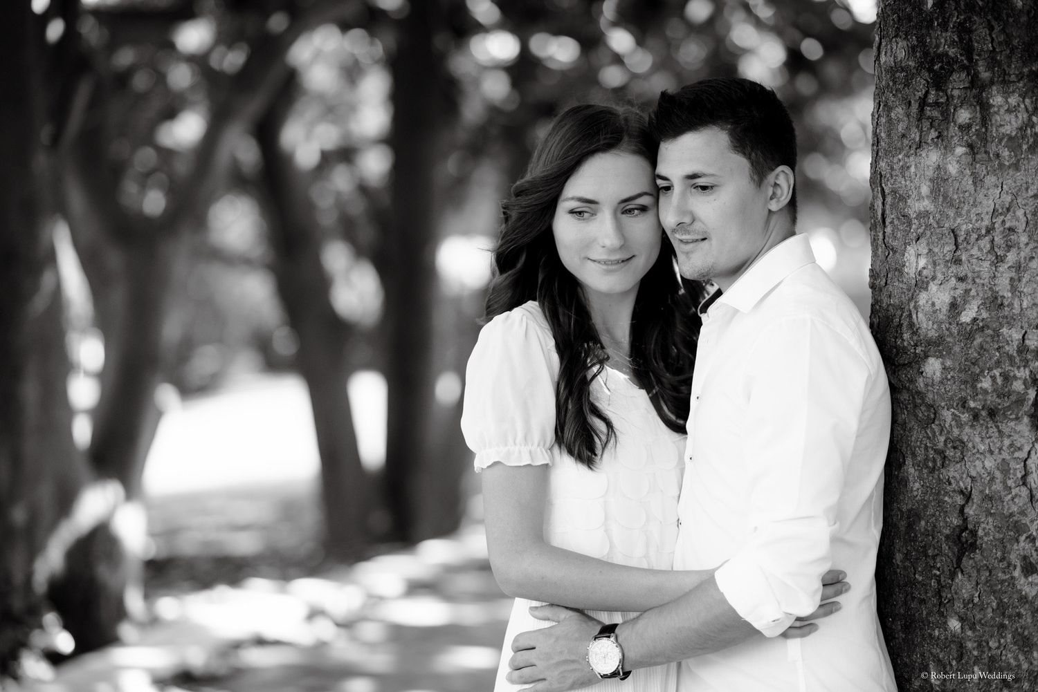 Black and white photo of a romantic couple embracing near a tree in a sunlit park, woman in white dress.