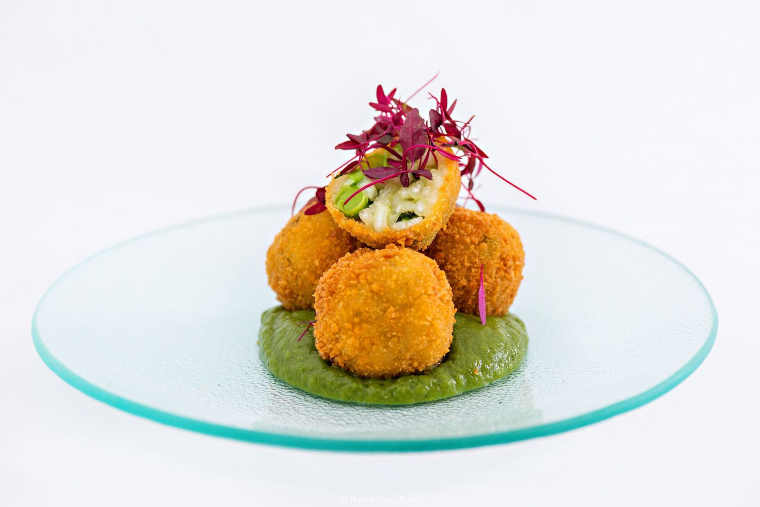 Golden fried croquettes on green puree, topped with fresh microgreens and sliced cucumber on a glass plate.