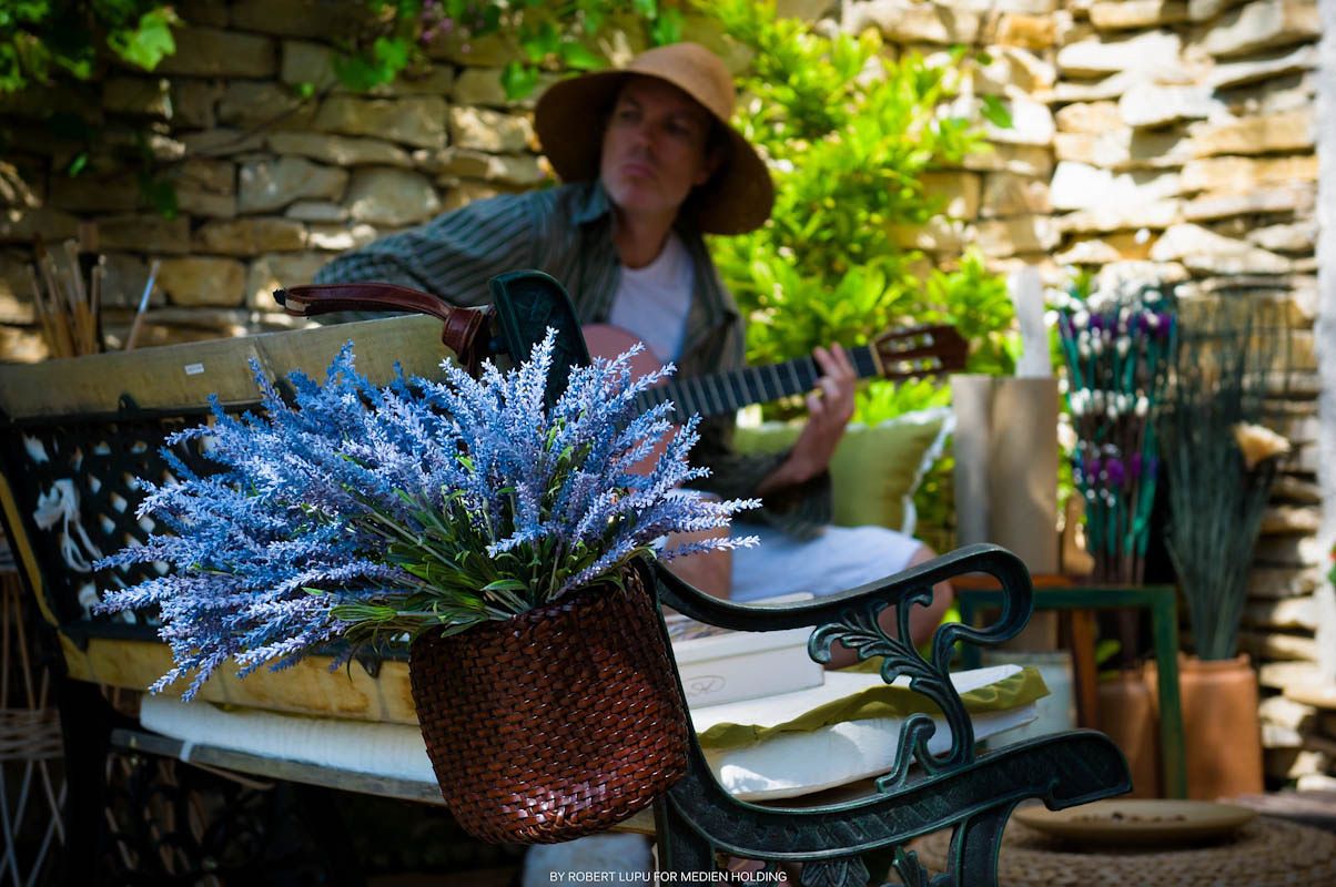 Woman in sun hat playing guitar outdoors beside a rustic stone wall with blue lavender flowers in a basket.