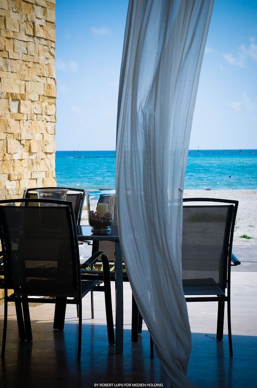 Beachfront patio dining area with black chairs, sheer white curtains, and stunning turquoise ocean views under a clear blue sky.