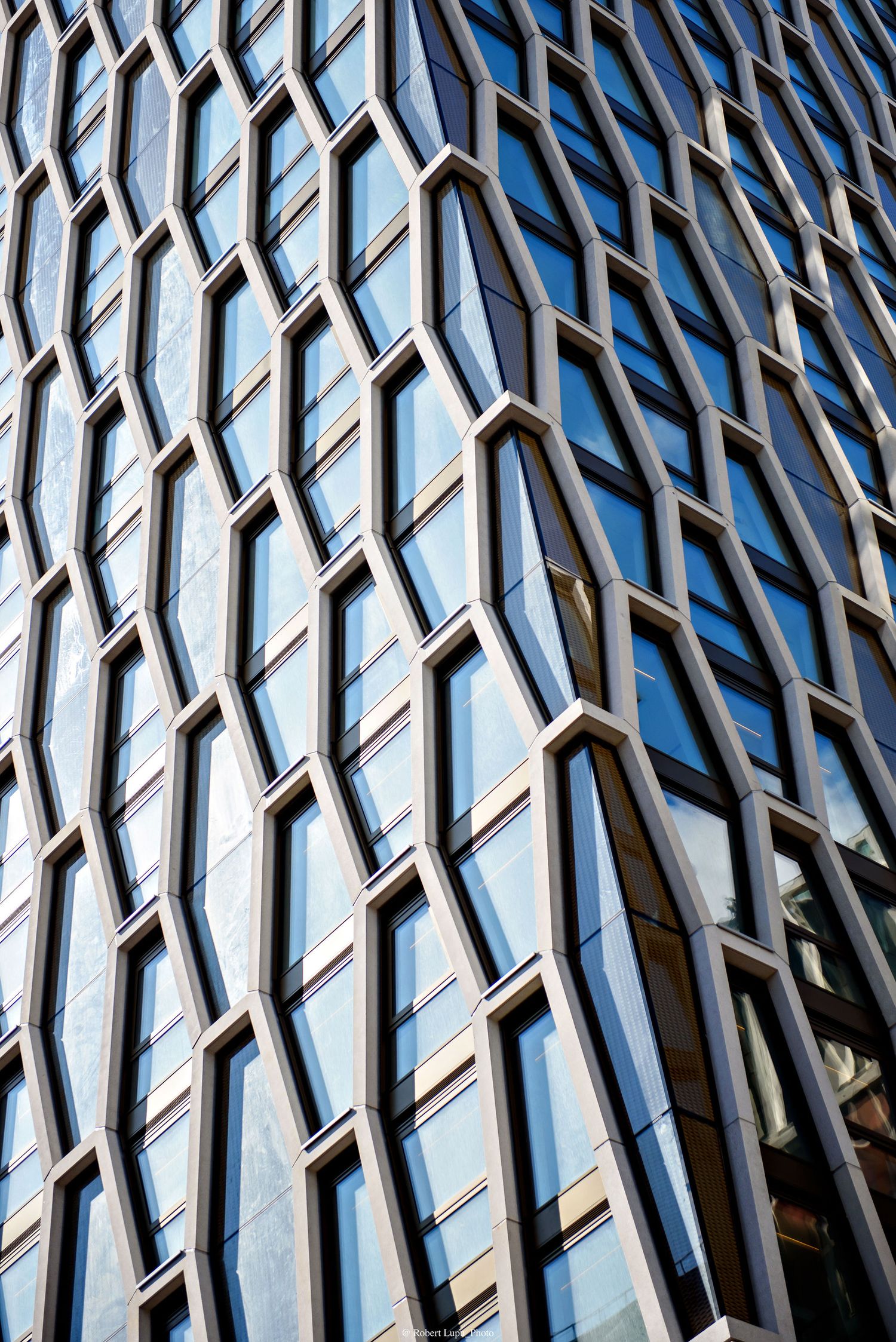 Modern glass skyscraper facade with geometric diamond-shaped window frames reflecting blue sky in an abstract architectural pattern.