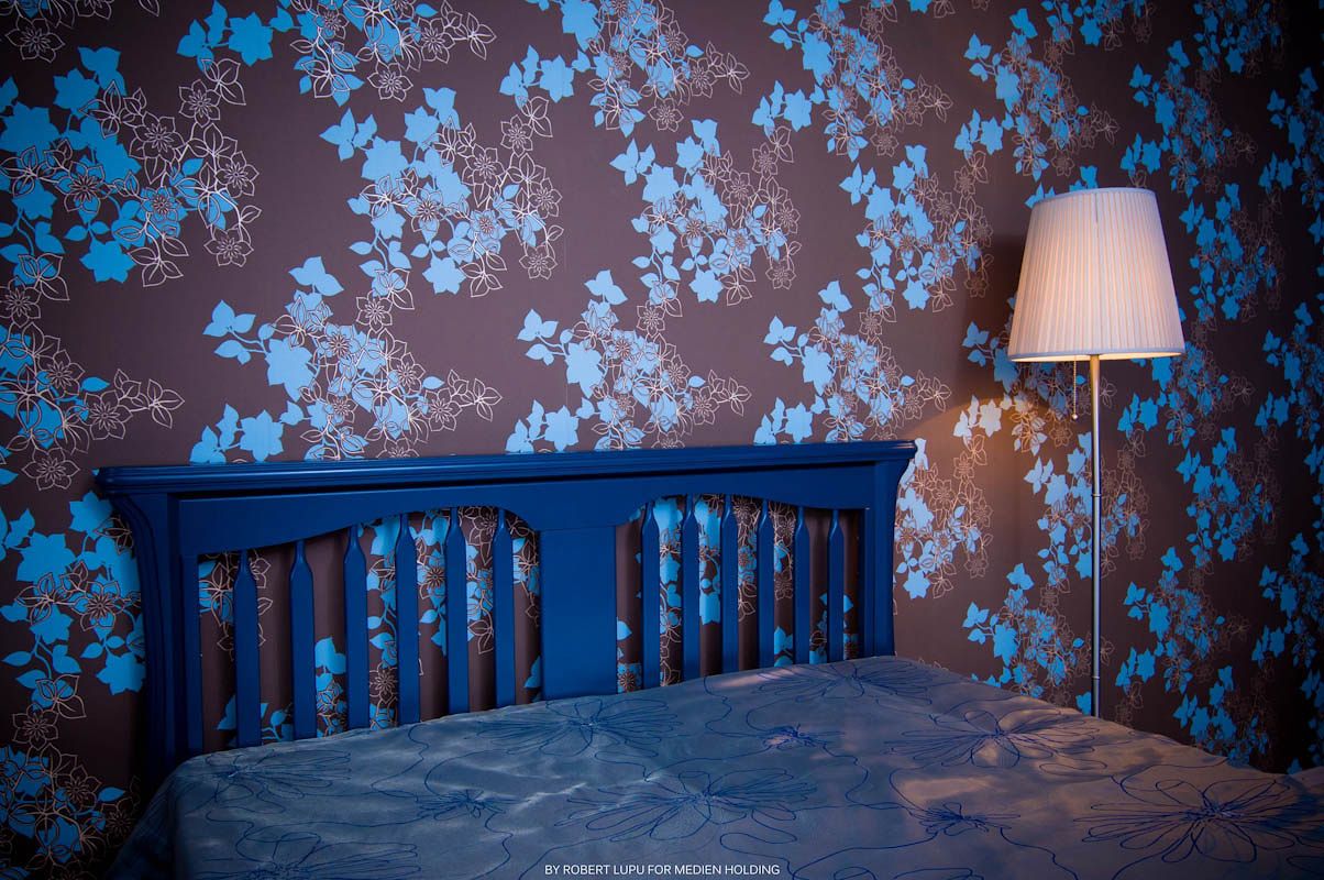 Blue floral wallpaper bedroom with navy wooden headboard and white floor lamp casting warm light.