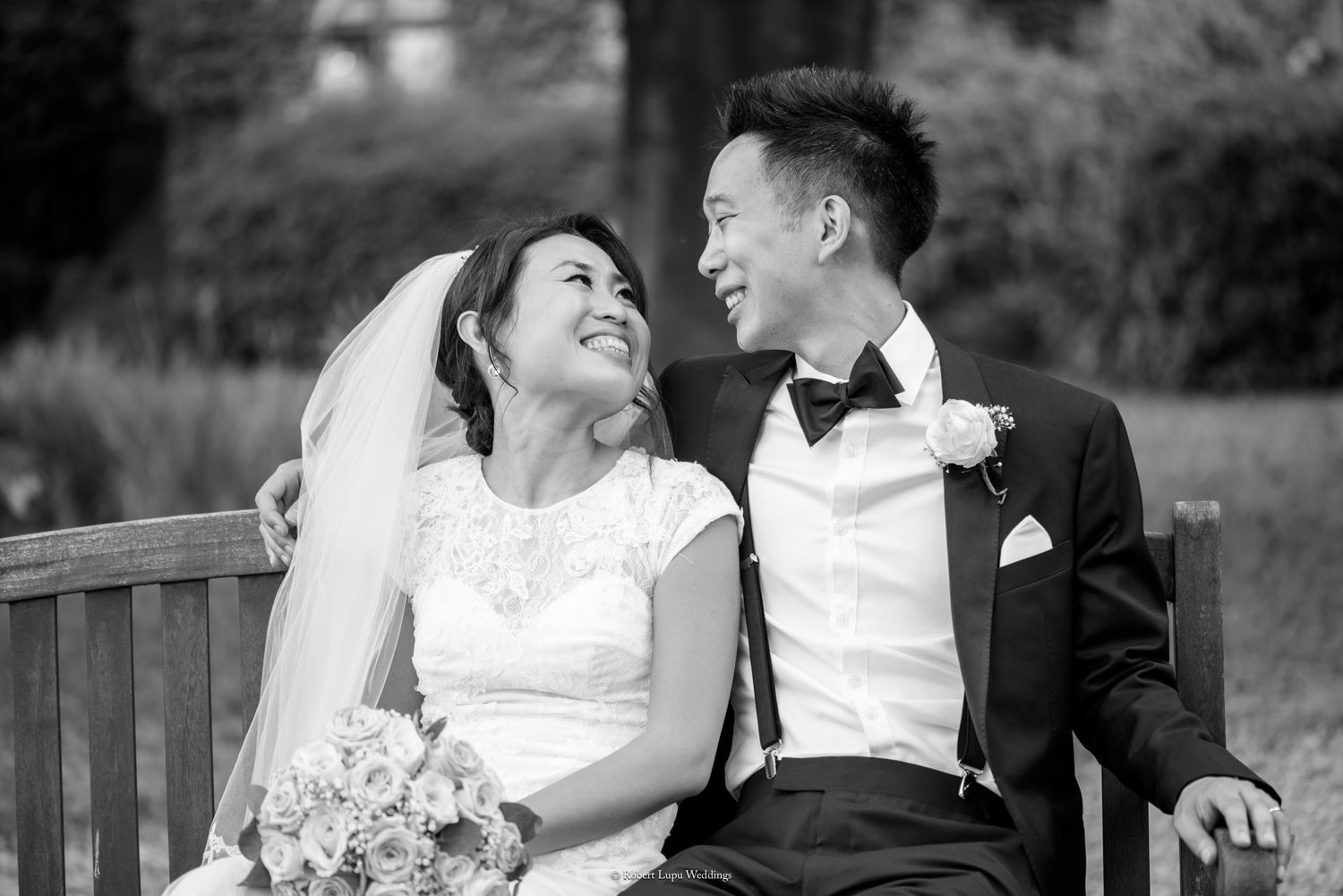 Black and white photo of a smiling Asian bride and groom sitting on a bench, sharing a loving glance on their wedding day.