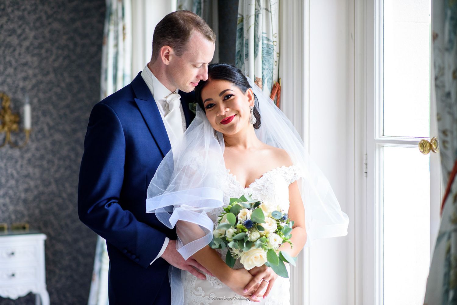 Elegant bride and groom posing together indoors, bride holding a white bouquet, groom in navy suit.