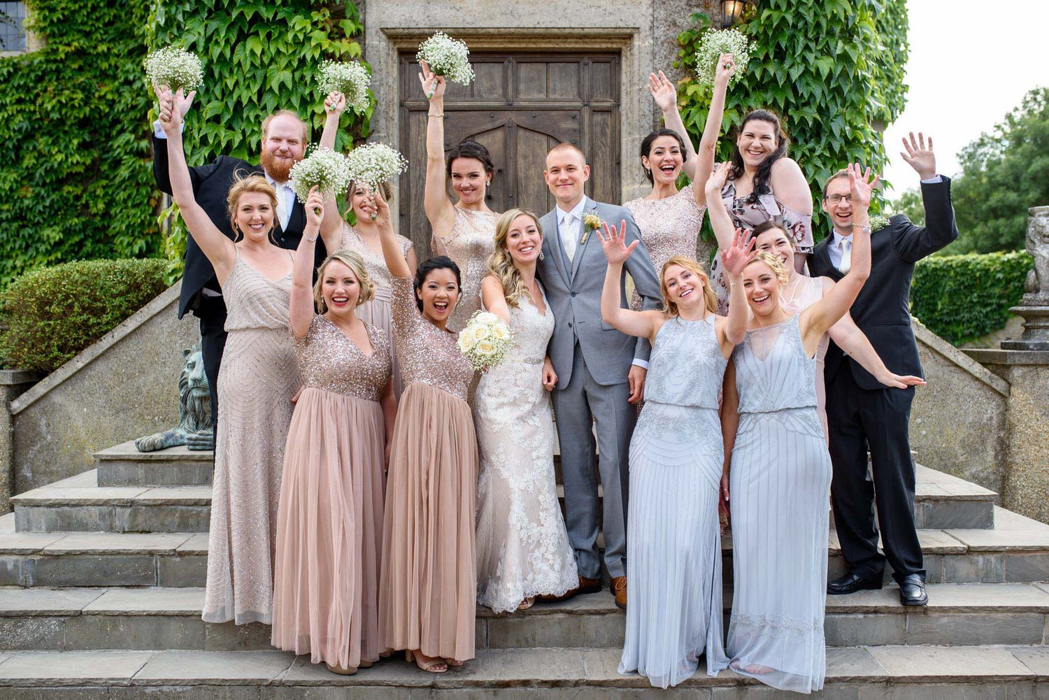 Wedding party poses on stone steps, bridesmaids in blush and blue gowns cheering with bouquets beside bride and groom.