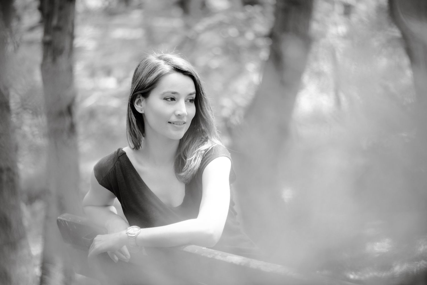Black and white portrait of a young woman smiling outdoors, leaning on a railing surrounded by trees.