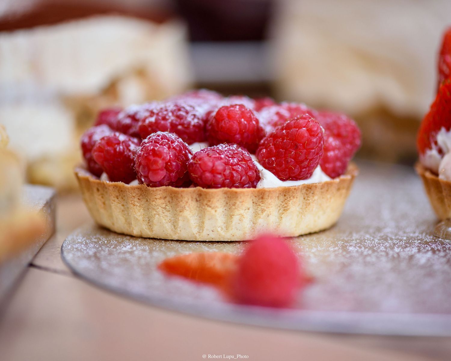 A golden pastry tart filled with cream and topped with fresh raspberries dusted with powdered sugar on a silver plate.