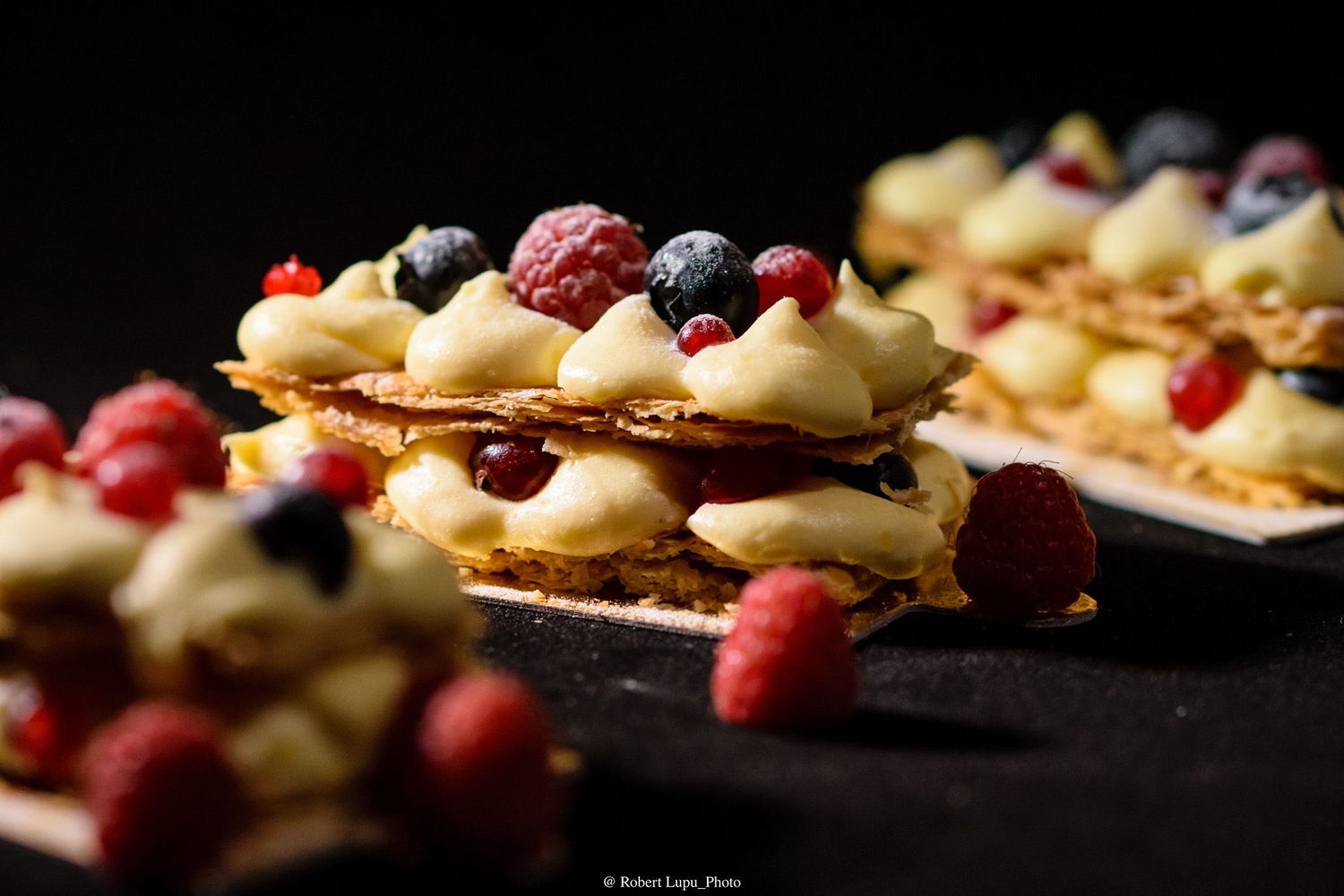 Elegant mille-feuille pastry layered with cream, fresh raspberries, and blueberries on dark background.