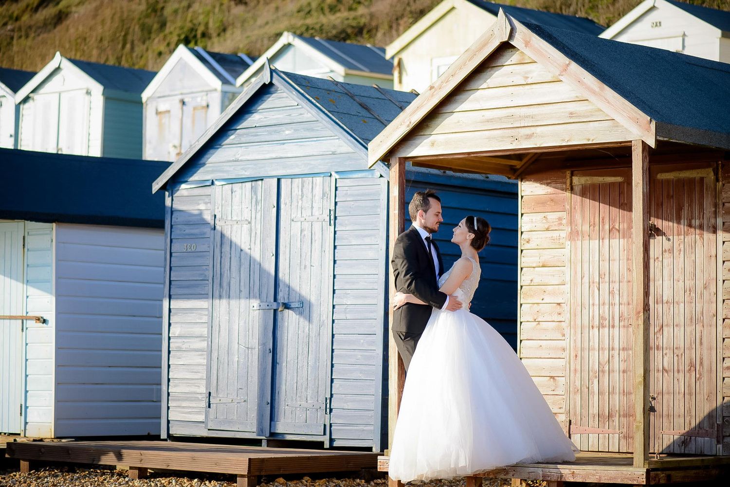 Bride and groom sharing a romantic moment between pastel beach huts on a sunny wedding day.