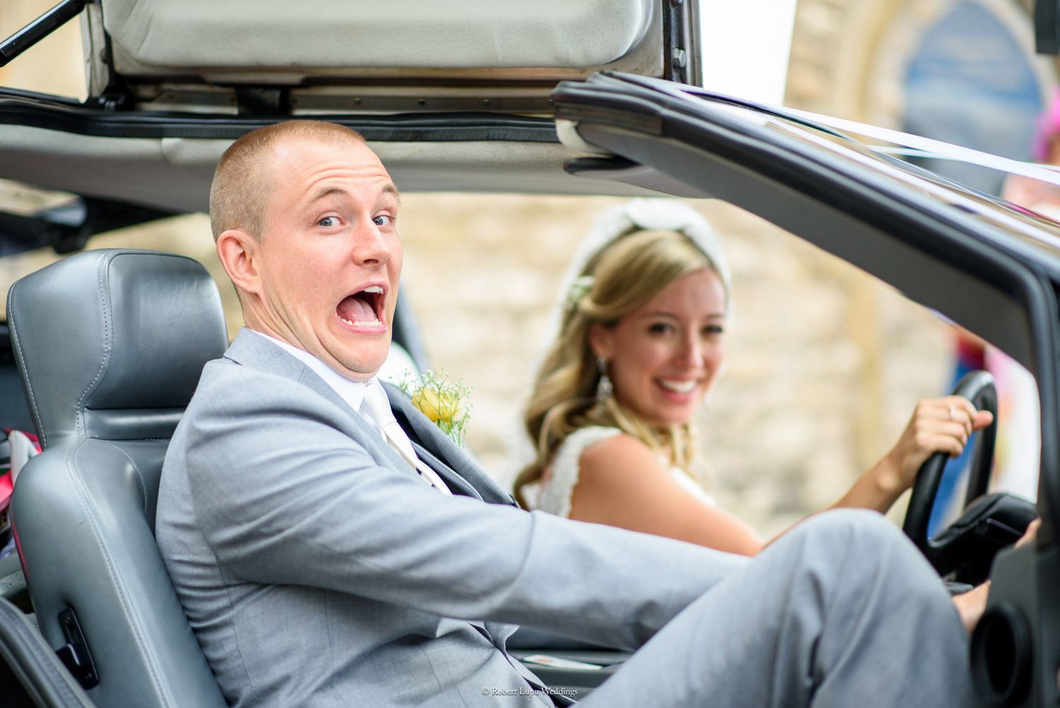 Surprised groom with open mouth sits in convertible car beside smiling bride in wedding attire.
