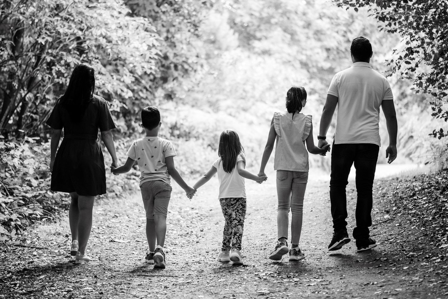 Black and white photo of a family of five holding hands walking down a tree-lined path together.