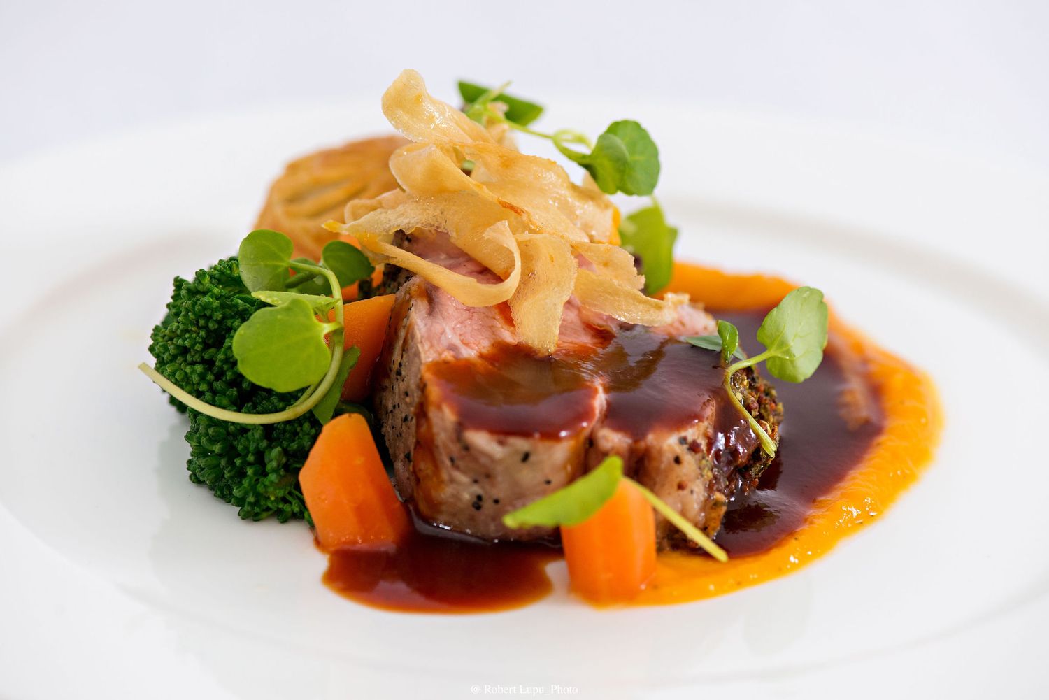 Elegant plated beef dish with broccoli, carrots, rich red wine sauce, and garnished with microgreens on a white plate.