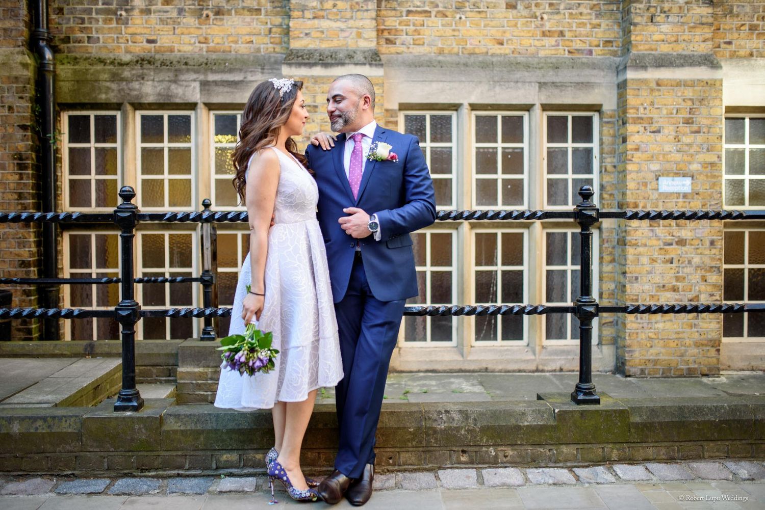 A bride and groom share a romantic moment outdoors, she holds a bouquet, he wears a navy suit with pink tie.