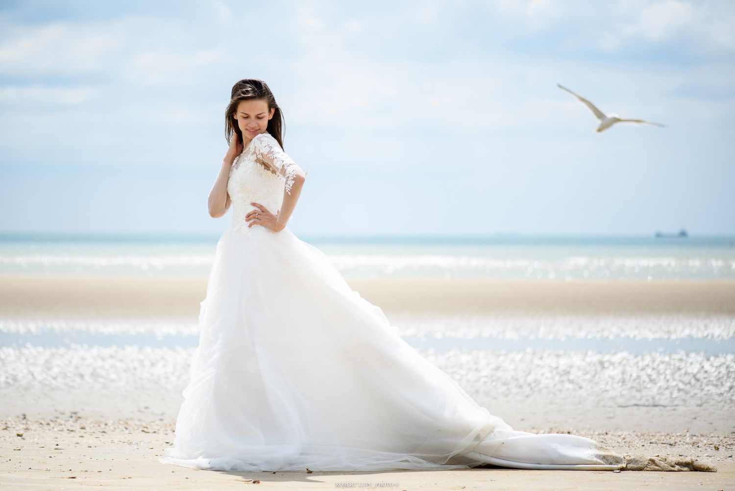 Elegant bride in flowing white ball gown poses gracefully on sunny beach with seagull soaring in background.