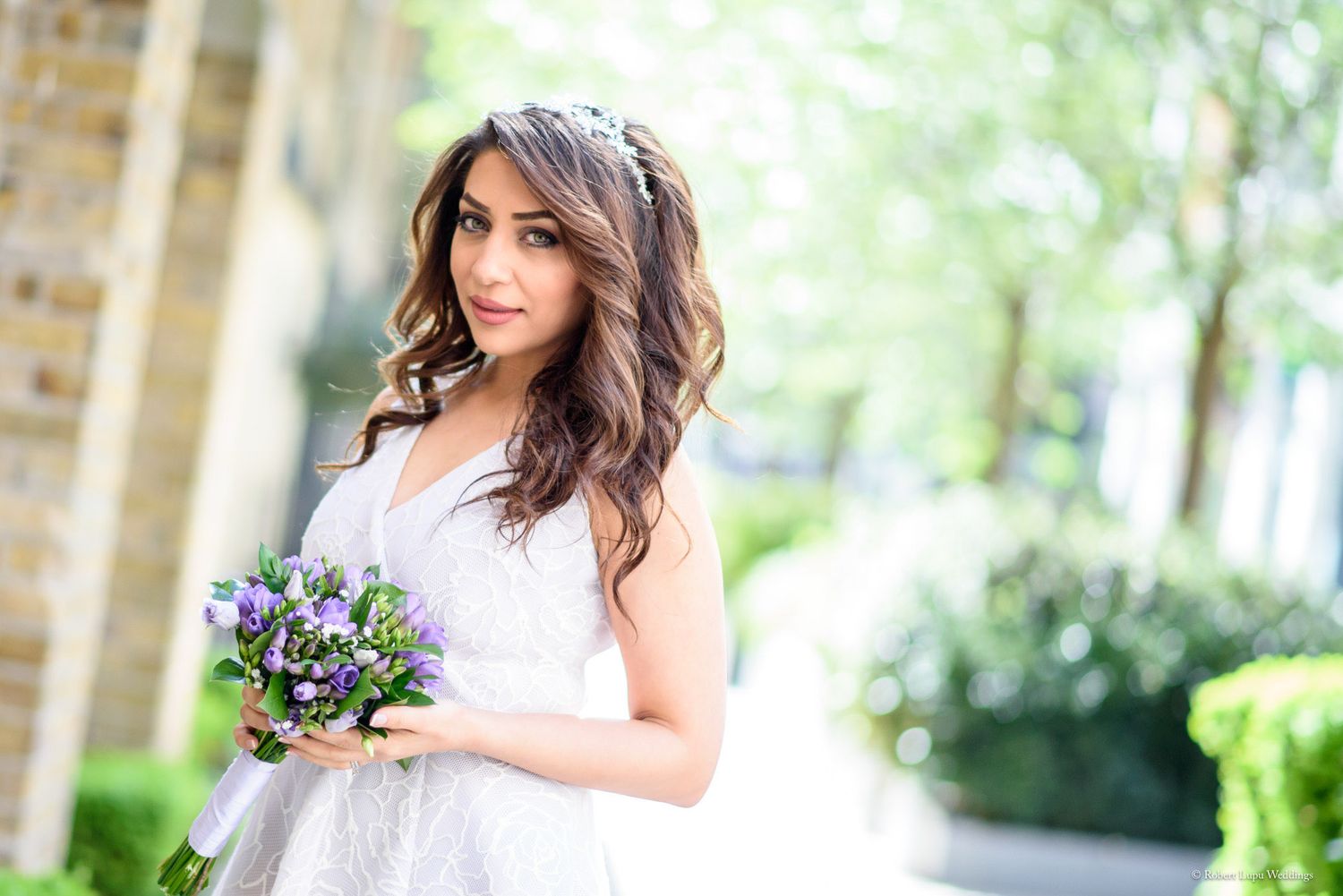 Bride in white lace dress holding purple flower bouquet outdoors.
