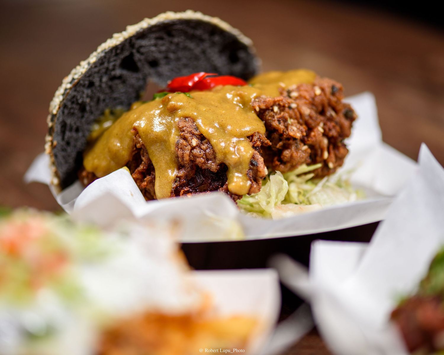 A gourmet black bun burger topped with melted cheese sauce and crispy fried chicken served in a paper-lined basket.