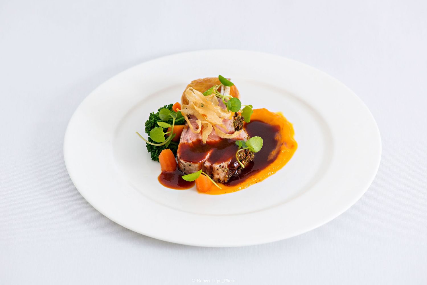 Elegant plated dish with seared meat, rich brown sauce, fresh greens, and carrots on a white plate.