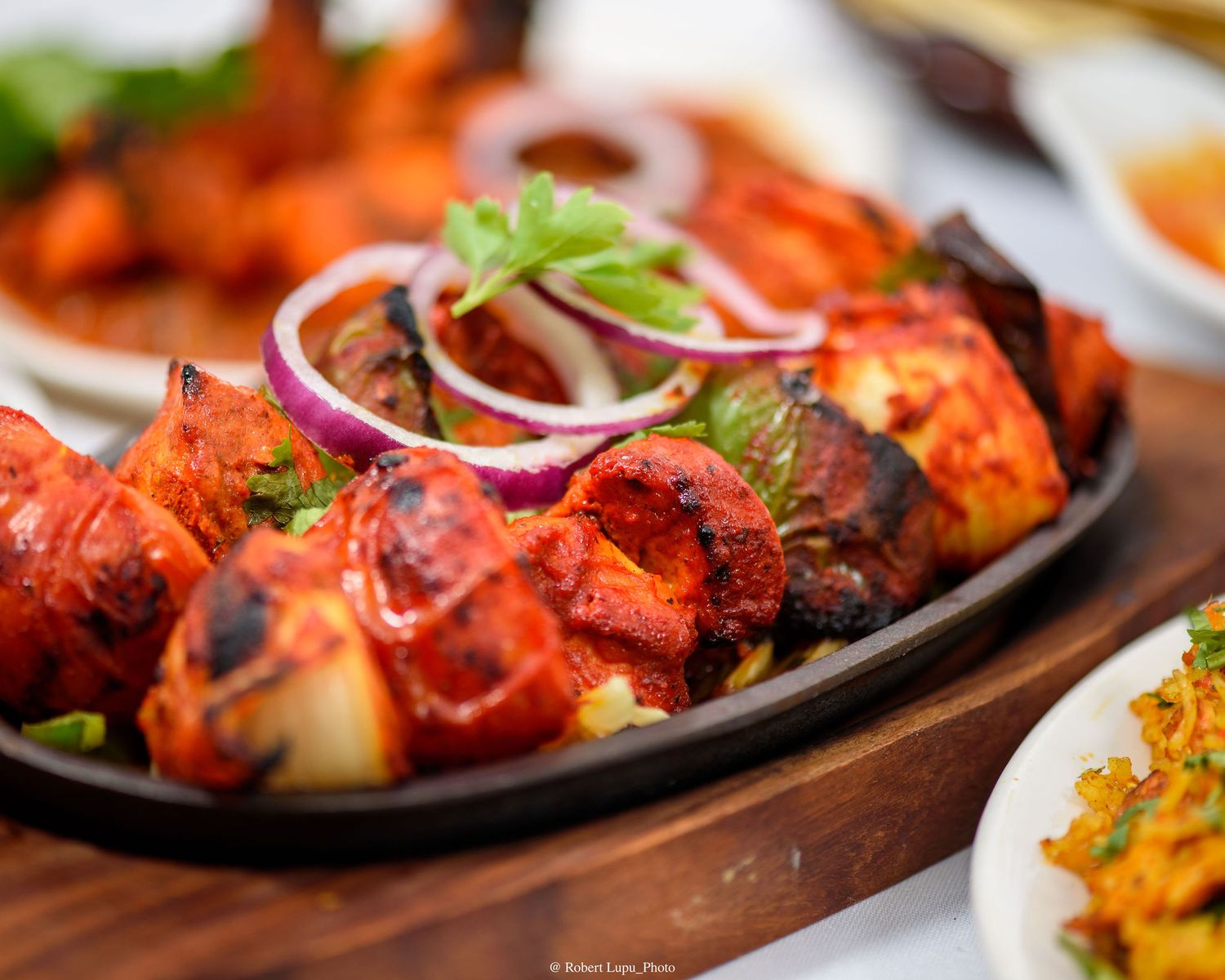 Tandoori chicken skewers on a sizzling platter, garnished with red onion rings and fresh cilantro.