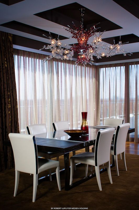 Modern dining room with white chairs, dark table, and red floral chandelier against sheer curtains.