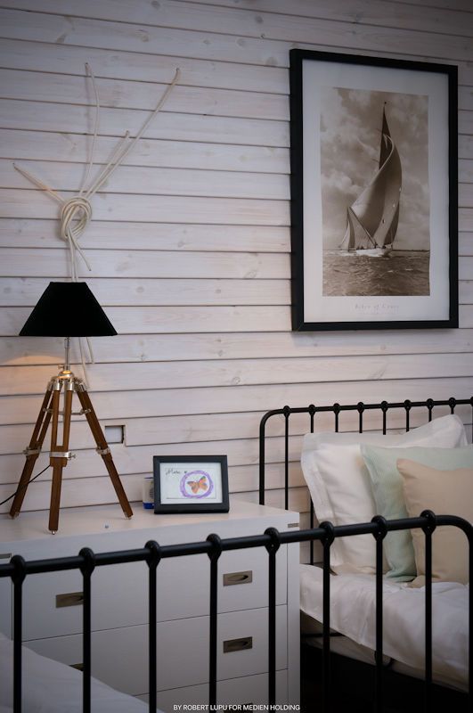 Nautical bedroom with iron bed, tripod lamp, sailboat artwork, and white shiplap walls for a coastal decor style.