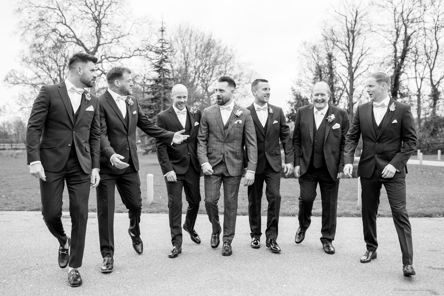 Groom and six groomsmen in dark suits walking together outdoors in a classic black and white photo.