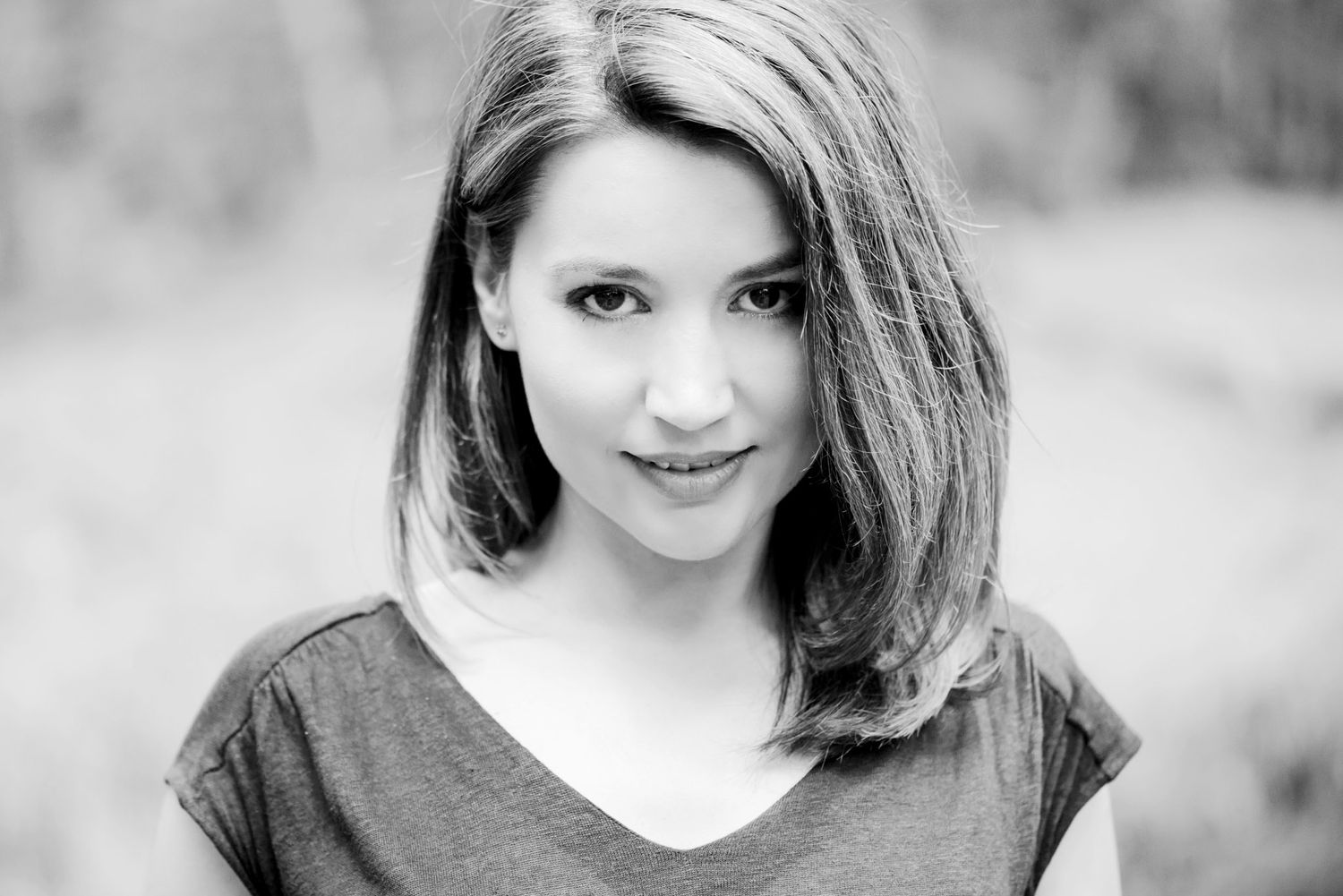 Black and white portrait of a young woman with shoulder-length hair wearing a v-neck t-shirt outdoors.