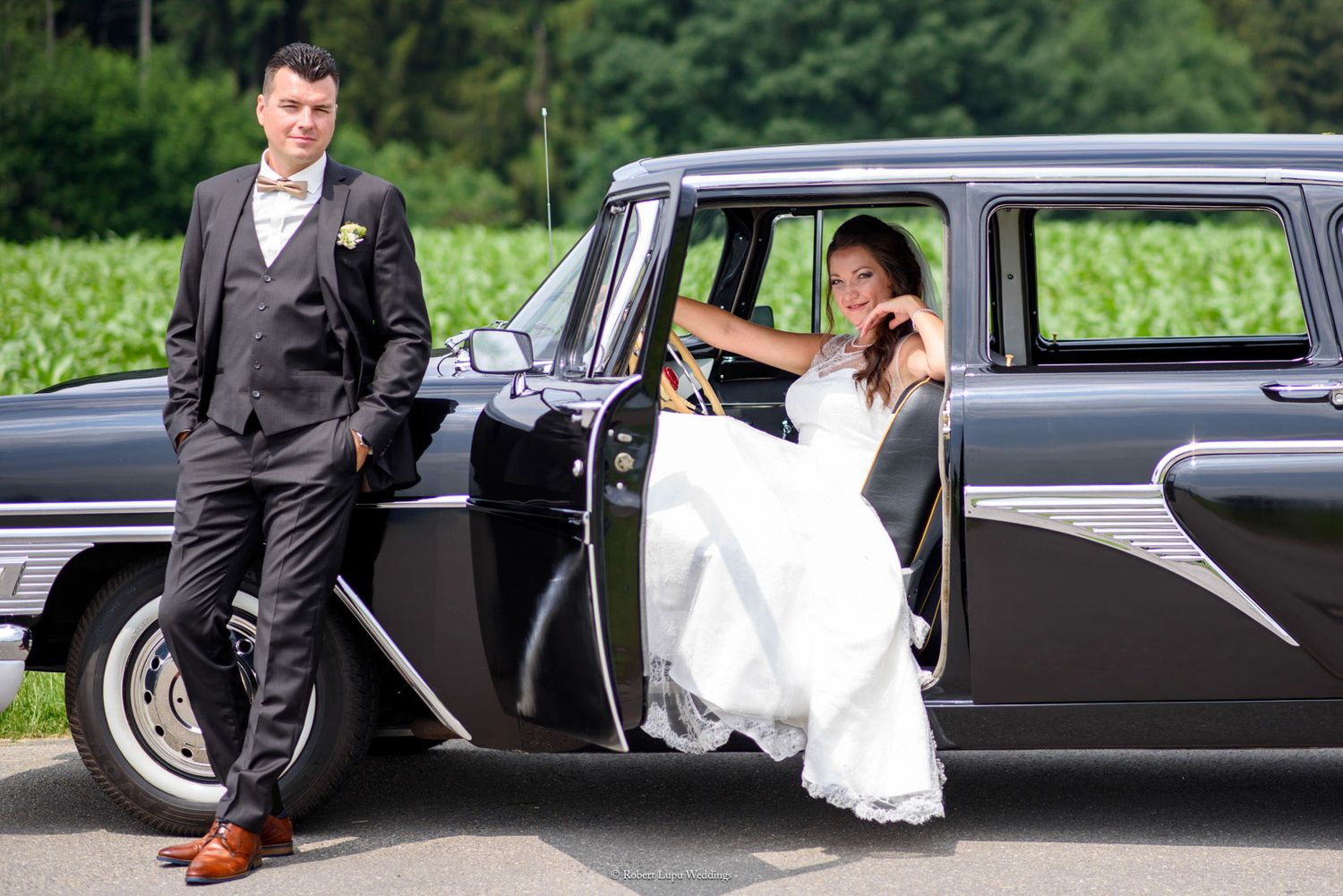 Bride in white gown sits in vintage black car as groom in dark suit stands beside it on their wedding day.