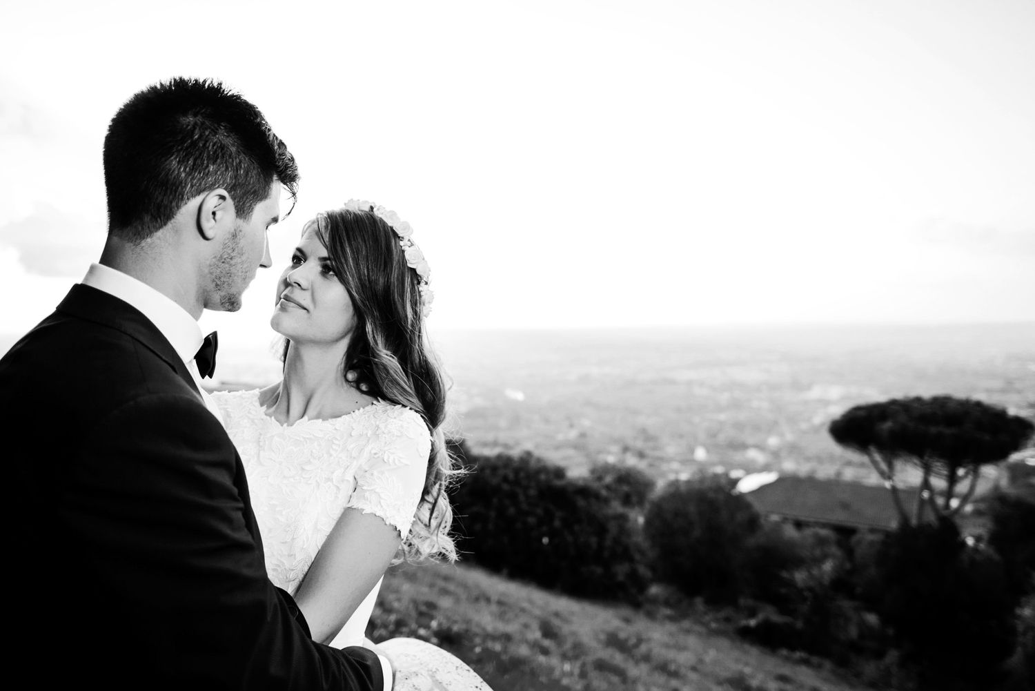 Black and white photo of a couple embracing on a hilltop with a scenic cityscape backdrop on their wedding day.