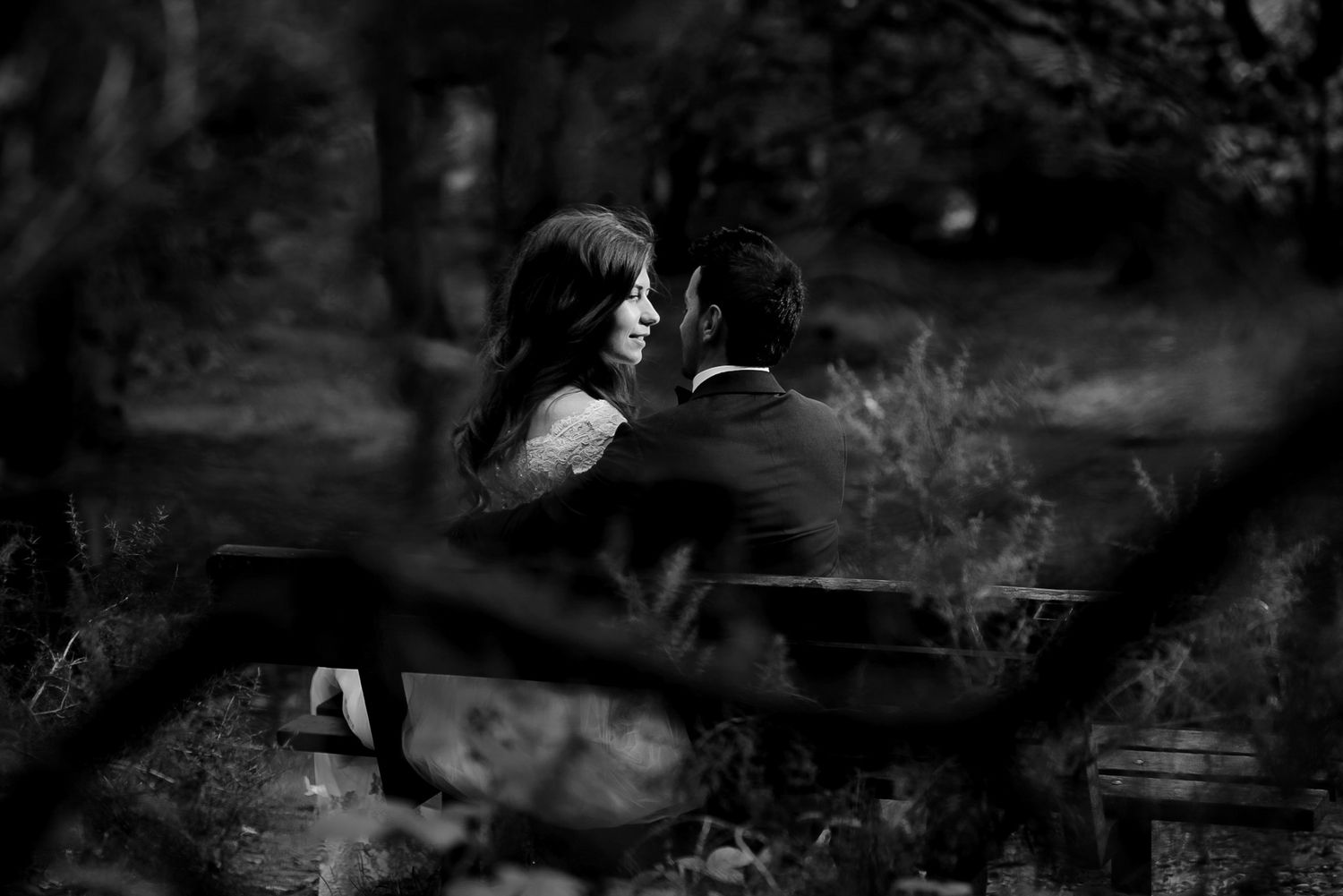Black and white photo of a couple sitting on a bench in a forest, gazing at each other romantically.