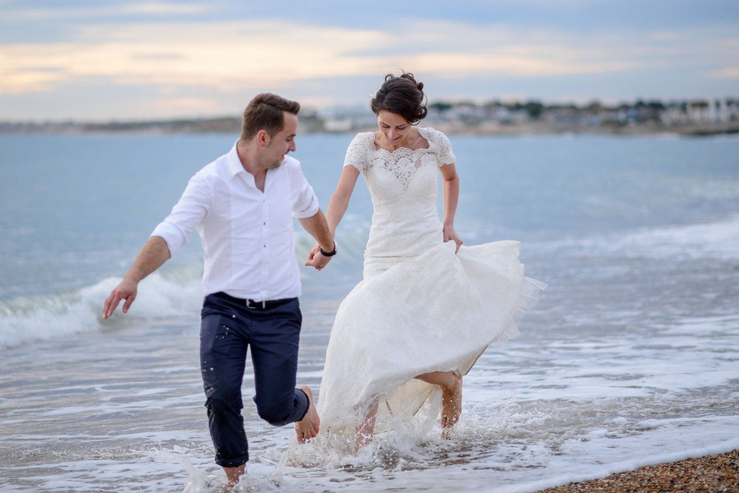 Newlywed couple running and splashing in ocean waves on a beach, bride in white gown holding hands with groom in white shirt.