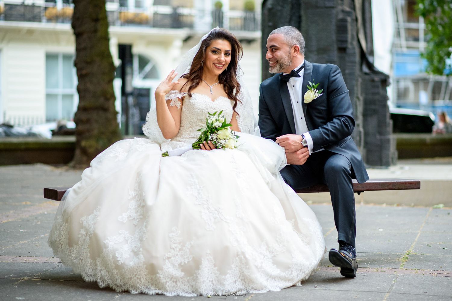 Bride in white ball gown and groom in tuxedo sitting on a bench outdoors on their wedding day.
